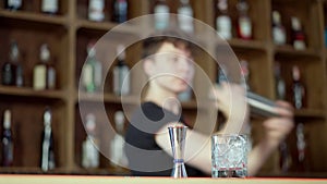 Jigger and glass with ice standing on bar counter with blurred young barman mixing drink in cocktail shaker at