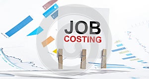 JOB COSTING text on paper sheet with chart, dice, spectacles, pen, laptop and blue and yellow push pin on wooden table - business