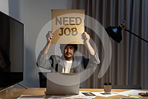 Job hunting. Man sitting at the table in the office, holding a piece of cardboard that says need job in his hands. Hire me concept