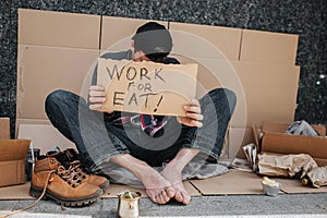 Jobless guy is sitting on the concrete ground and covering his face with sign work for eat. He is looking for a job. Guy