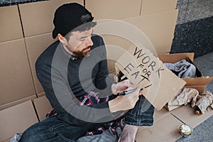 Jobless man is sitting on the ground and writing down words work for eat on the piece of paper. Guy is looking for a job