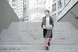 Jobless woman goes down the stairs on the street with a box of personal items. Female employee in medical mask fired due