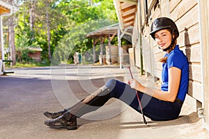 Jockey girl sitting outside a box stall with whip