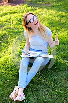 Jocund female student showing thumbs up studying in park and sitting on green grass.