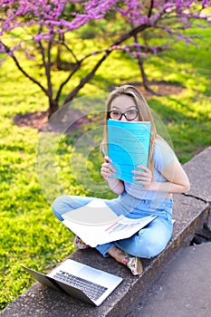 Jocund female student sitting with laptop and papers in folder in blooming park.