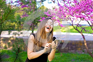 Jocund girl using new smartphone in park with blooming trees background.