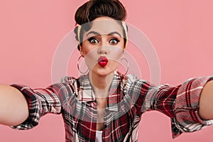 Jocund pinup girl posing in red checkered shirt. Amazed woman taking selfie with kissing face expression