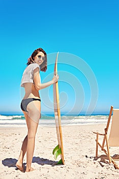Join me for a surf. Rearview portrait of a sexy young woman standing on the beach with her surfboard.