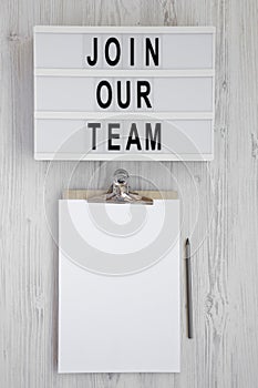 `Join our team` words on a lightbox, clipboard with blank sheet of paper on a white wooden background. Overhead, from above, fla