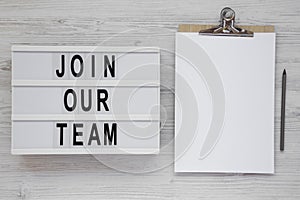 `Join our team` words on a modern board, clipboard with blank sheet of paper on a white wooden background. Overhead, from above,
