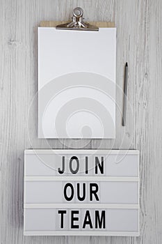 `Join our team` words on a modern board, clipboard with blank sheet of paper on a white wooden background, top view. Overhead,