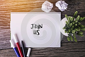 Join Us word with Notepad and green plant on wooden background.