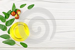 Jojoba oil in a bowl, a sprig of jojoba and fruits on a wooden table