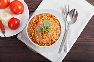 Jollof rice, tomatoes and hot peppers on a blue plate, fork, spoon on a linen napkin on a brown wooden background. National