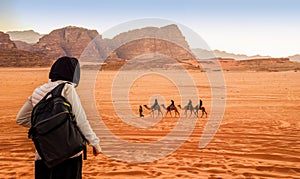 Jordan, Wadi Rum. Caravan of camels with drovers in the desert, rock mountains at sunset. Tourist from behind looks at the