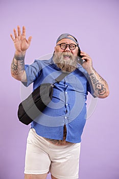 Jouful plump man talks by phone and waves hand standing on purple background photo