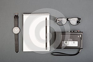 Journalist or blogger table - spiral blank notebook, pencil, tape recorder and glasses on gray background, top view