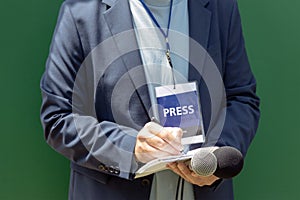 Journalist with press pass at news conference or media event, holding microphone, writing notes