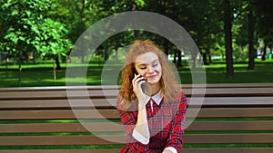Jovial red haired girl talking on smartphone in park and laughing
