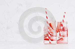 Joy bright fresh summer drinks with red strawberry, straw, ice, soda water in elegant jars on soft light white wood table.