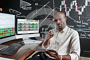 Joy of earning money. Cheerful african american male trader sitting by desk, pointing at camera and studying analytical
