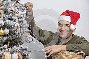 A joyful 30-35-year-old man in a red hat is decorating a Christmas tree with a garland and lights, looking into the camera.