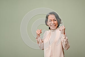 Joyful 60s aged-asian woman raises hands with clenched fists, standing against green background
