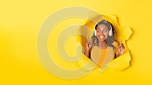 Joyful african american lady listening favourite music in wireless headphones, posing in paper hole on yellow background