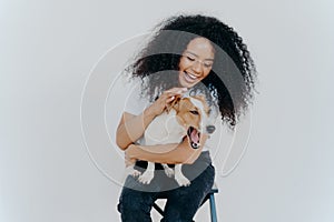 Joyful African American woman plays with pedigree dog, dressed in casual wear, petting favourite pet, isolated over white