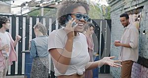 Joyful African American woman speaking on mobile phone laughing standing in street cafe during open air party