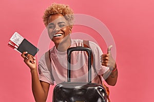 Joyful african american woman with tickets, passport and suitcase on pink background.