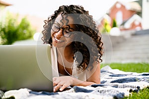 Joyful african american woman using laptop while lying on blanket on grass
