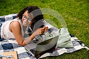 Joyful african american woman using laptop while lying on blanket in green park