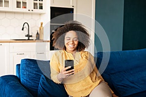 Joyful African-American woman using smartphone sitting on the couch