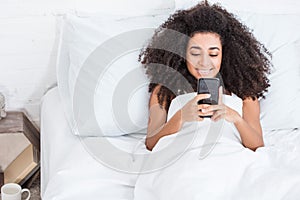 Joyful african american young woman using smartphone in bed during morning time