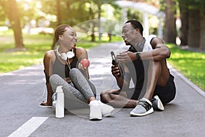 Joyful african couple resting after exercising outdoors, sitting on path in park
