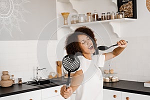 Joyful African young woman having fun with kitchen utensils. African american housewife in apron singing with spatulas