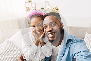 Joyful afro father and daughter wearing crowns and taking selfie