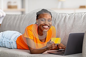 Joyful Afro woman shopping online with laptop pc and credit card, lying on couch at home