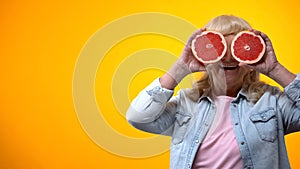 Joyful aged lady holding two slices of grapefruit on orange background, vitality