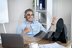 Joyful Arab company employee listening to music in headphones and playing virtual guitar at desk in modern office