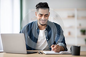 Joyful arab man taking notes while working with laptop
