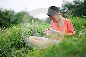 Joyful asian boy student sitting writing and reading book outside on school or park.