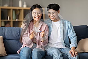 Joyful Asian Couple Holding Positive Pregnancy Test Sitting At Home