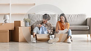 Joyful Asian couple sitting on floor of new house, unpacking boxes, relocating to their own property, moving in together