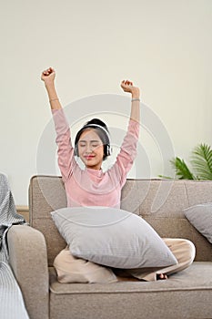 Joyful Asian girl listening music on headphones, raising hands and relaxing on her sofa