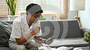 Joyful asian man using smart phone while sitting on couch in living room.