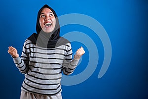 Joyful asian student girl in headscarf celebrating success with raised hands and clenched fists