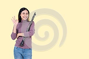 A joyful Asian woman showing the Okay hand sign and holding a clarinet, isolated yellow background
