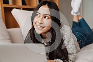 Joyful asian woman smiling and using laptop while lying on couch